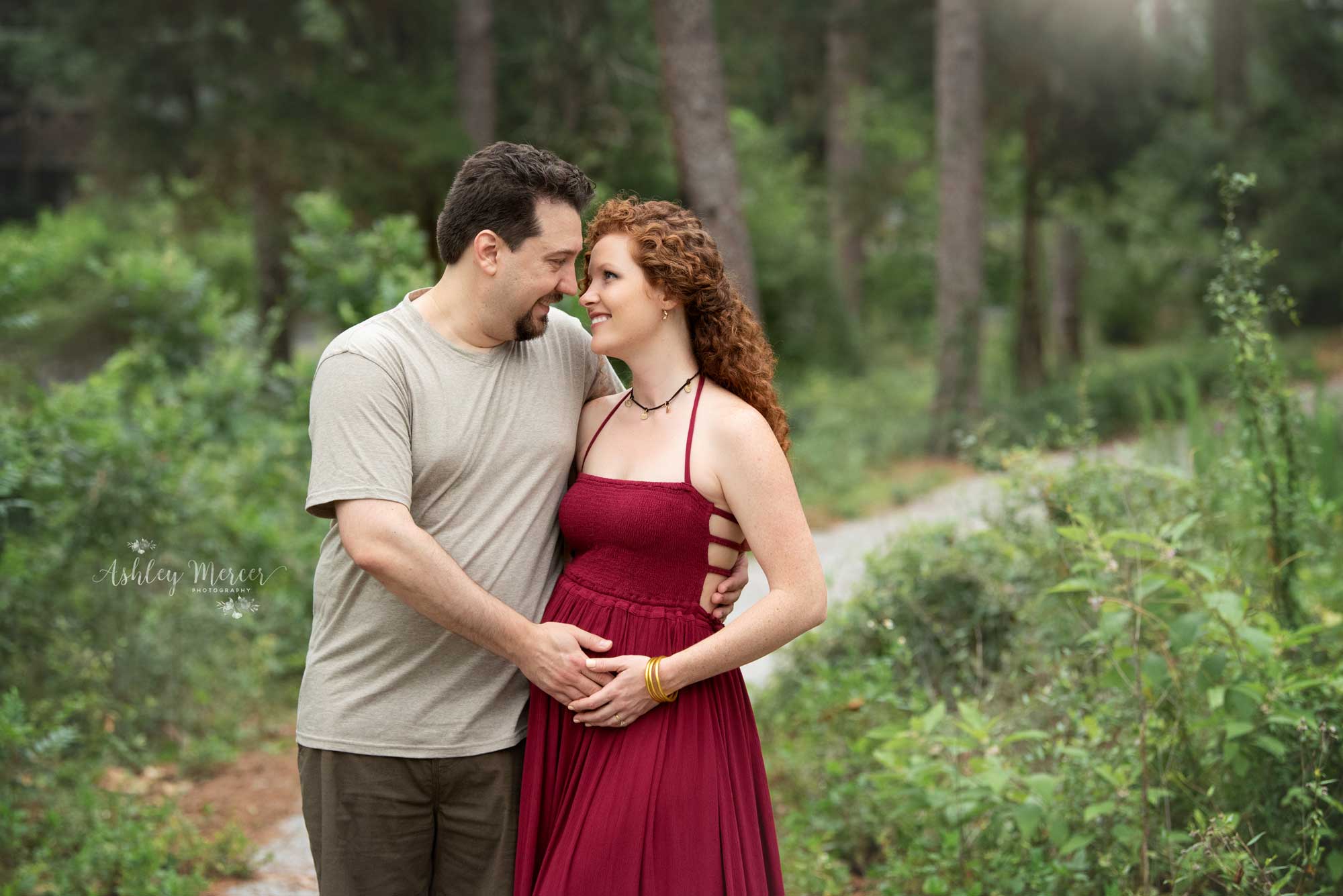 An expecting couple is holding each other while on a trail in the woods.