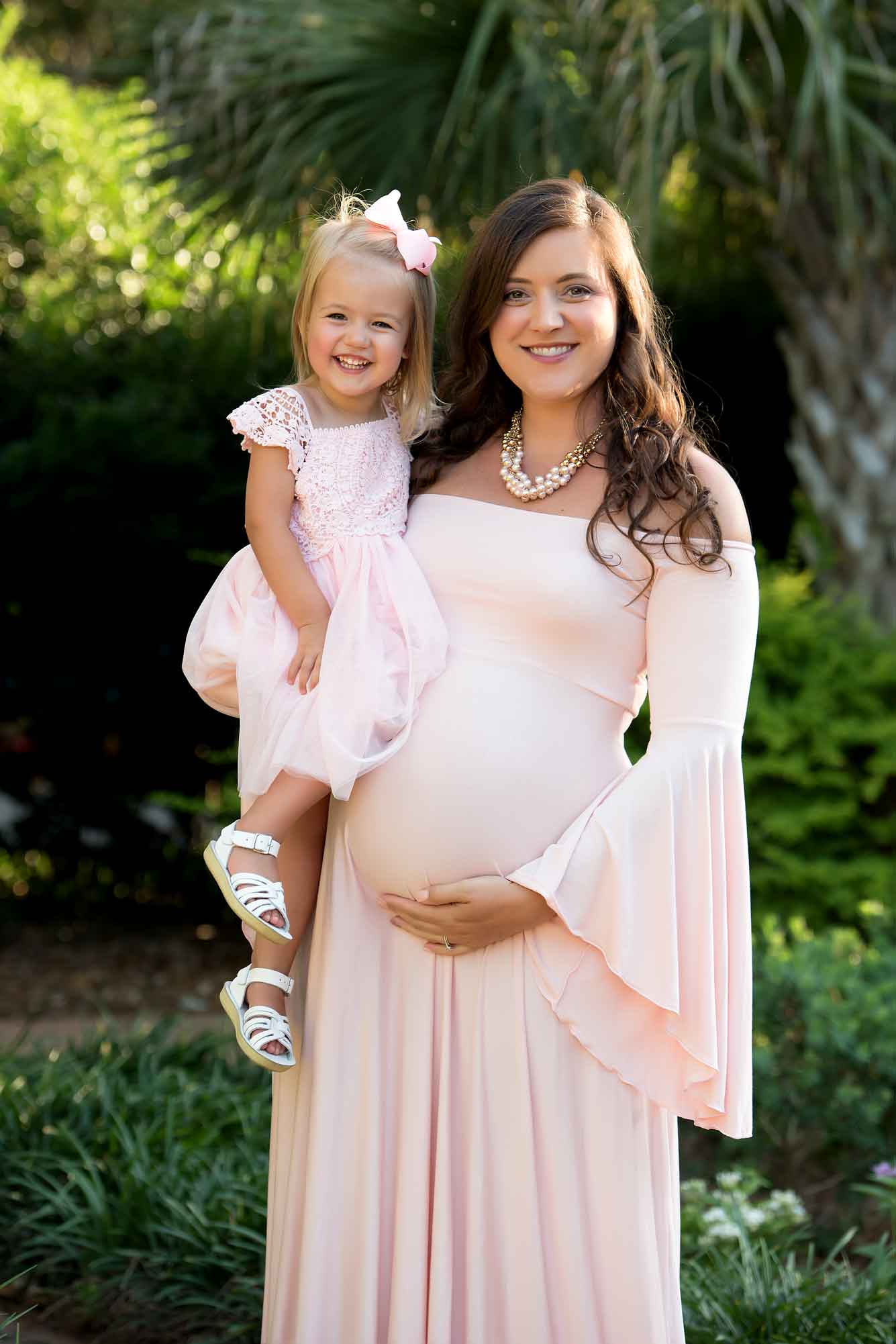 A mom and daughter, in matching pink dresses smile at the camera in green garden. The mom is holding the little girl while also showing off her baby bump