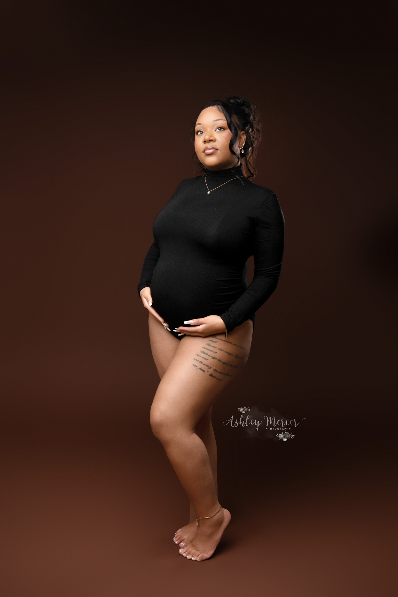 A fun and edgy maternity session with the mom wearing a black, long sleeve fitted body suit. She has her hands on her stomach and is looking off into the distance. The backdrop is dark brown giving the image a moody feel
