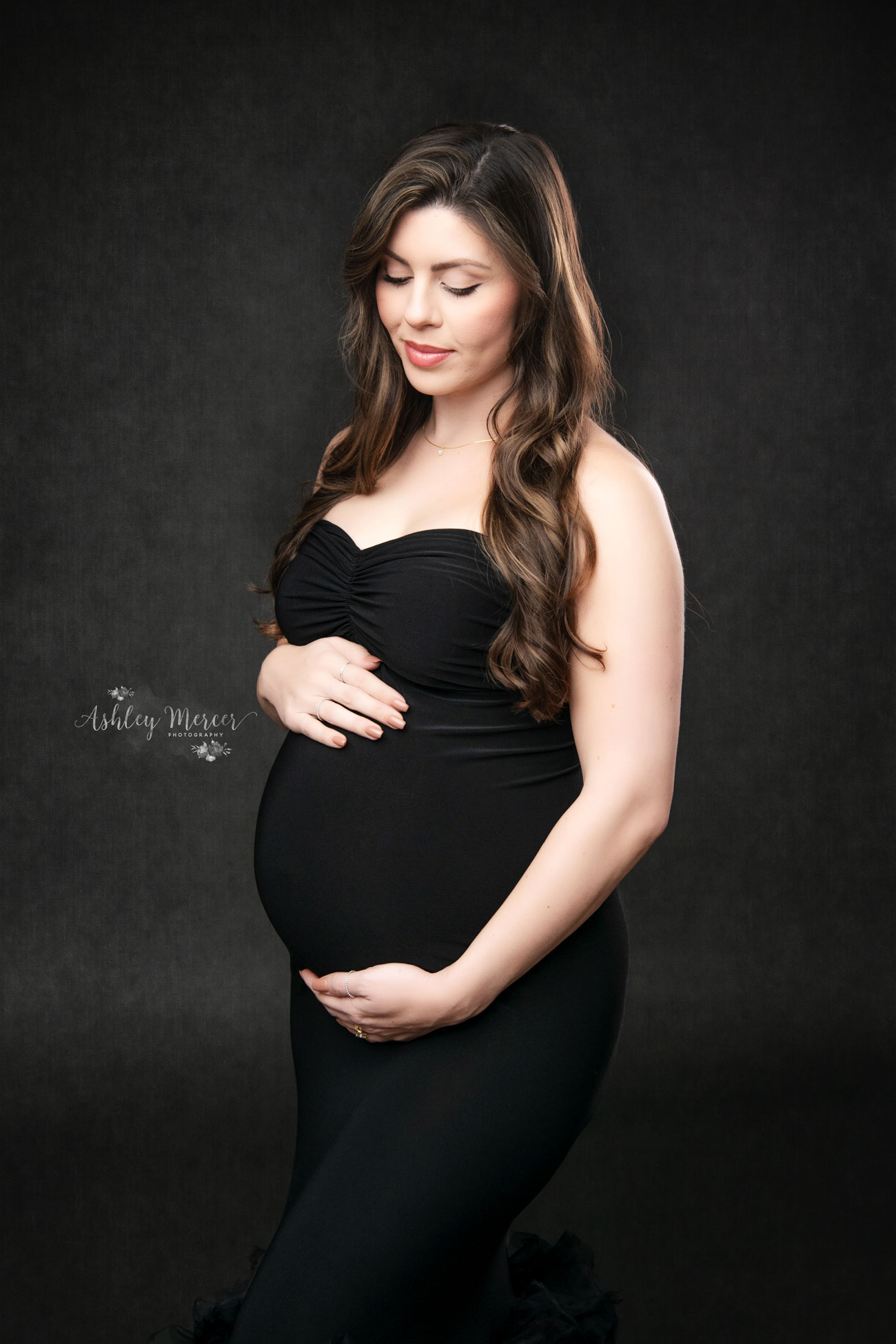 An expectant mother lovingly looking down at her belly while wearing a fitted back gown in the studio set against a dark grey backdrop