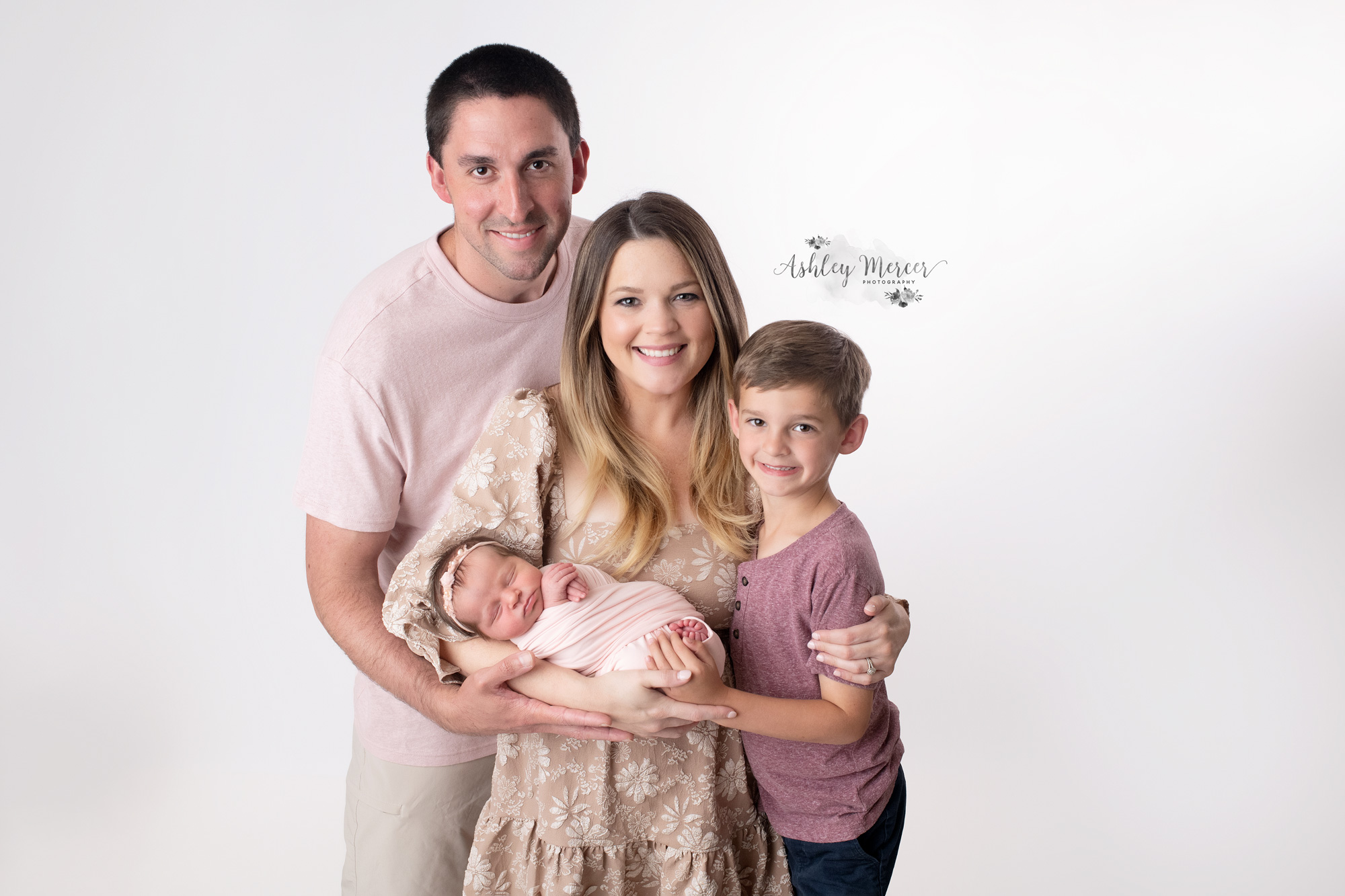 Family newborn portrait of a mom, dad, big brother, and baby sister. They are against a bright white background and the father is hugging everyone
