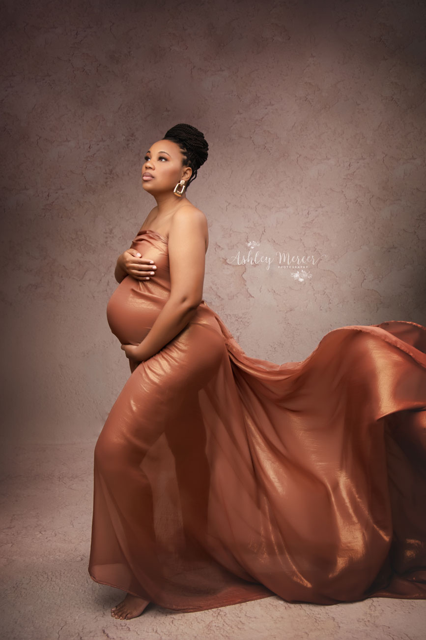 A mother to be is standing with her profile to the camera holding her belly. The train of her gown is flowing behind her