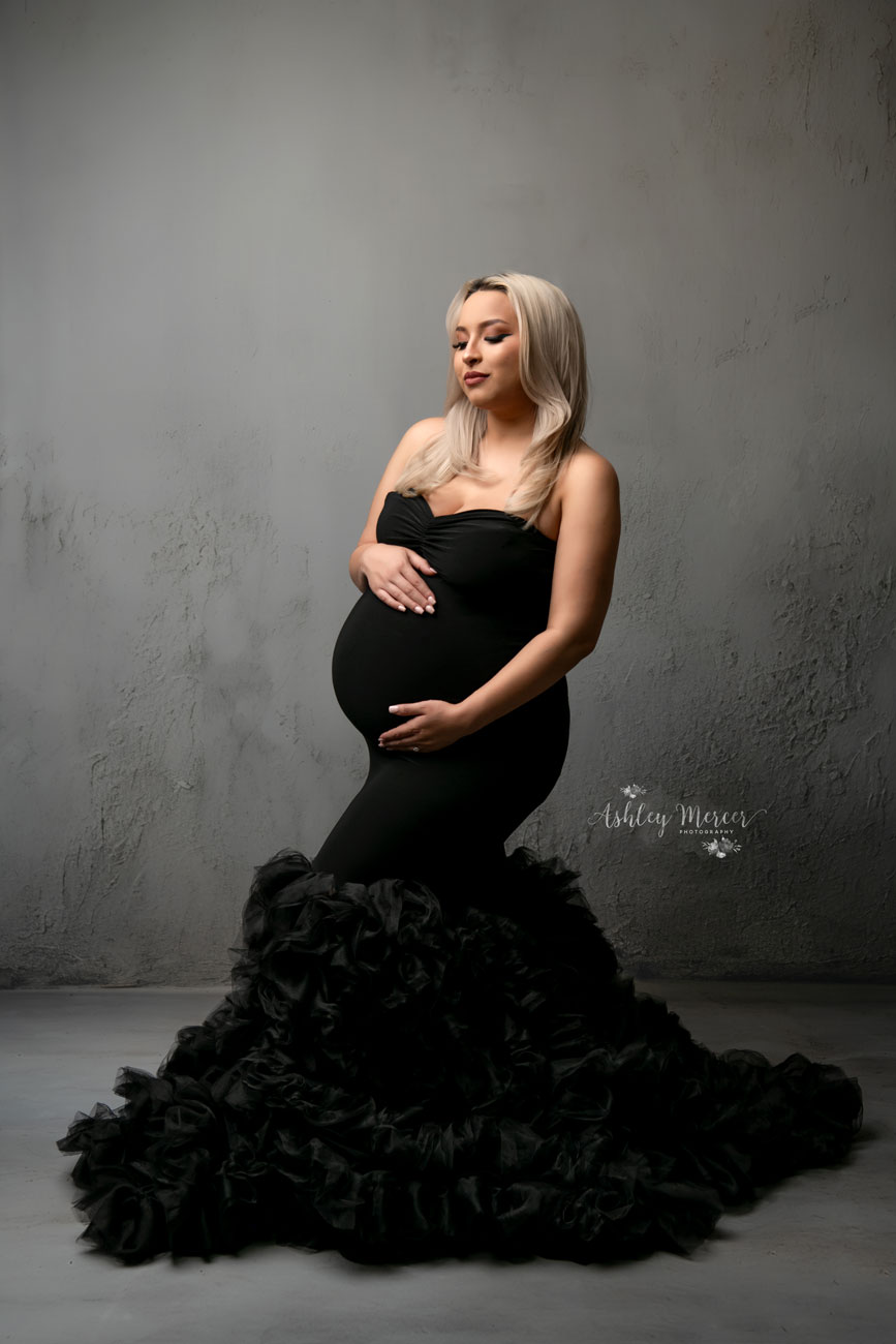 An edgy photoshoot with a pregnant mom wearing a fitted black gown that flares wide at the bottom. The lighting is moody and she is against a dark grey background