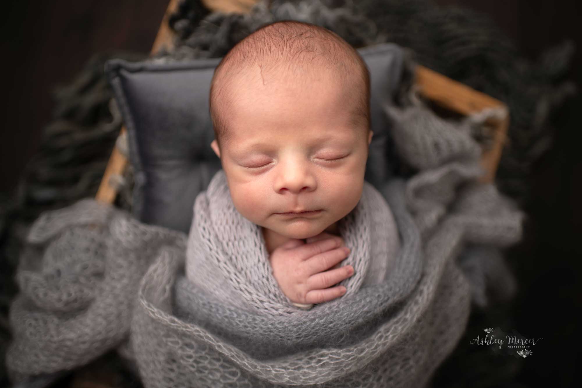 Baby boy wrapped in several grey knit wraps with both hands by his chin. He is propped up in a crate with more grey textures underneath
