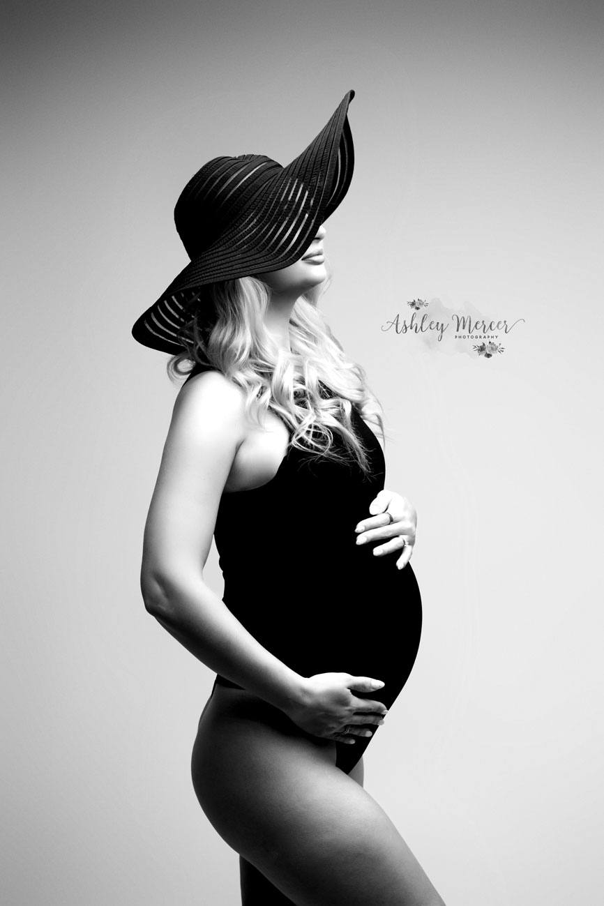 A black and white maternity portrait of a woman wearing a large, dramatic floppy hat while cradling her belly