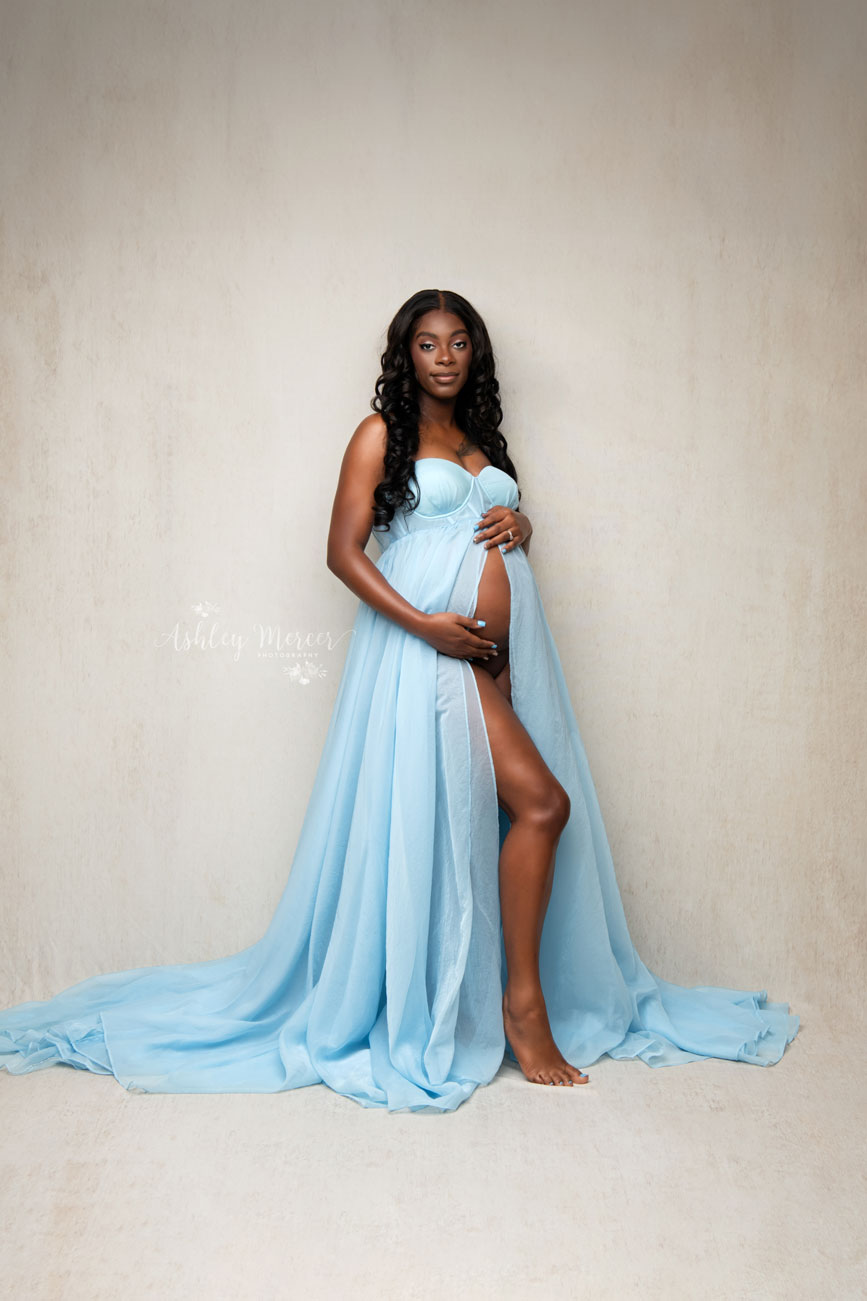 A pregnant woman wearing a blue maternity gown, showing of her baby bump. Bright and airy lighting gives it a classic feel.
