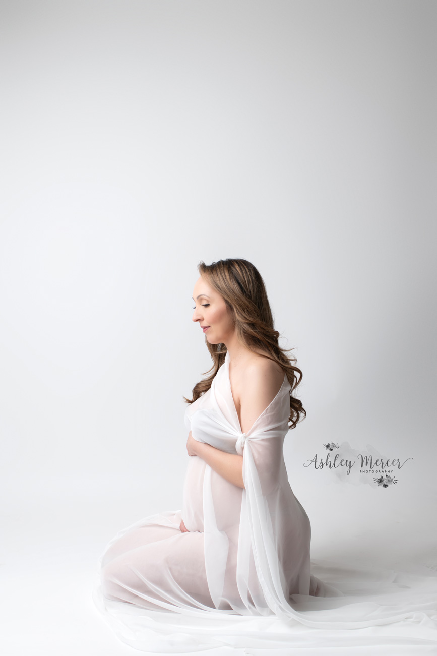 A romantic portrait of a pregnant woman sitting on her feet looking lovingly at her belly. She is draped in white sheer fabric