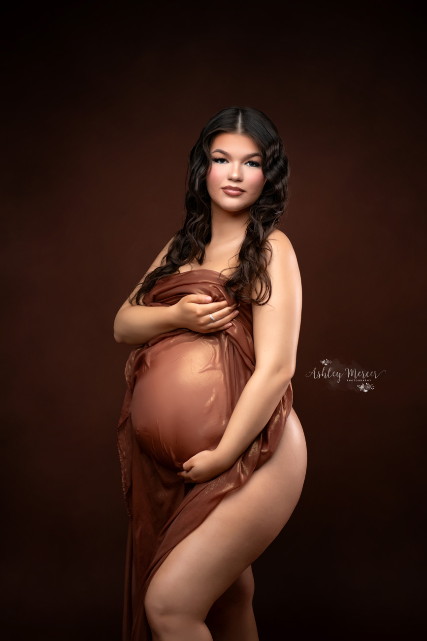 The sexy maternity portrait has a dark brown background with moody lighting. The mom is draped in sheer fabric, while cradling her baby bump and looking at the camera