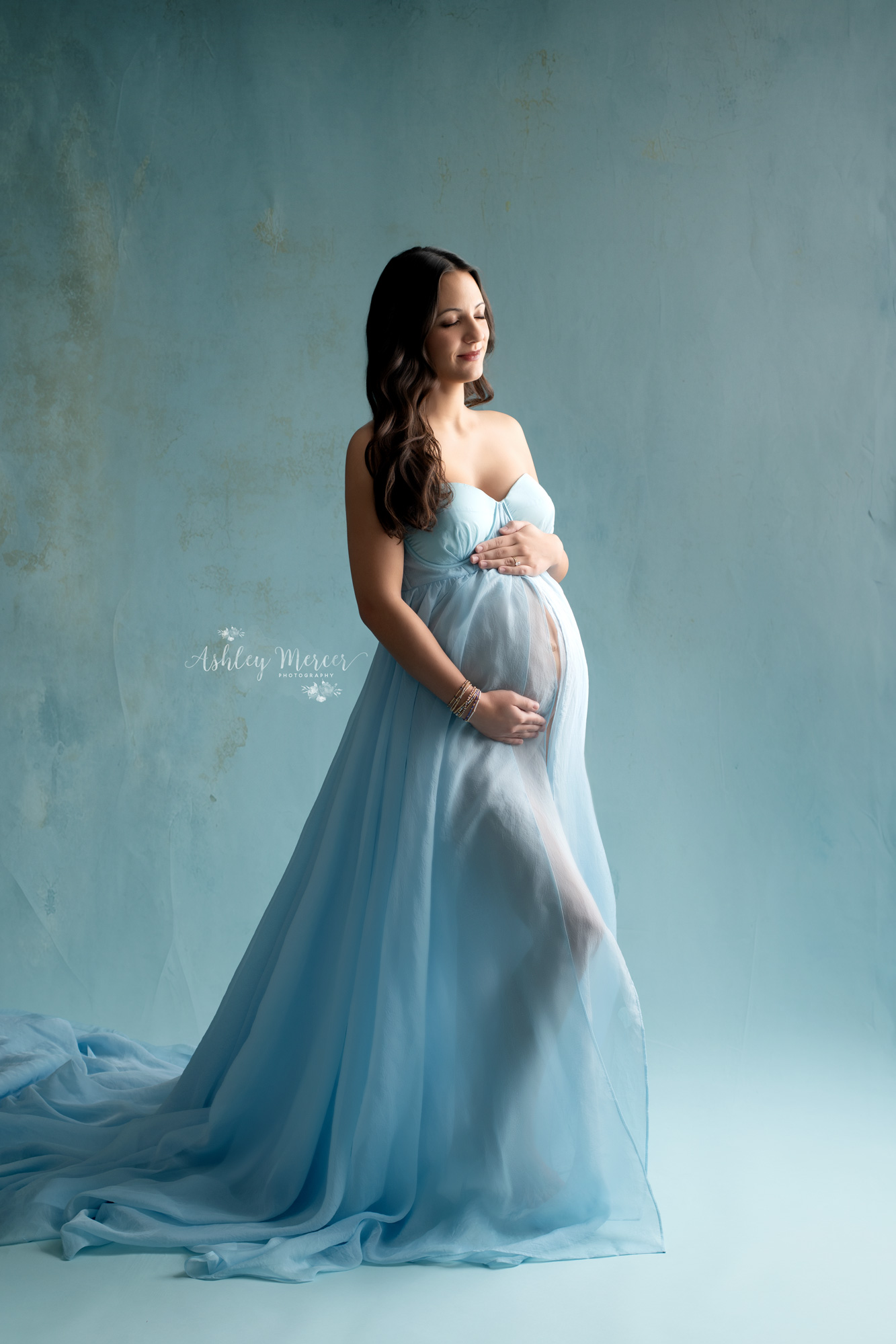 A mother to be is lovingly holding her belly and looking at it. She is wearing a romantic blue, flowing gown with dreamy lighting accentuating her growing belly