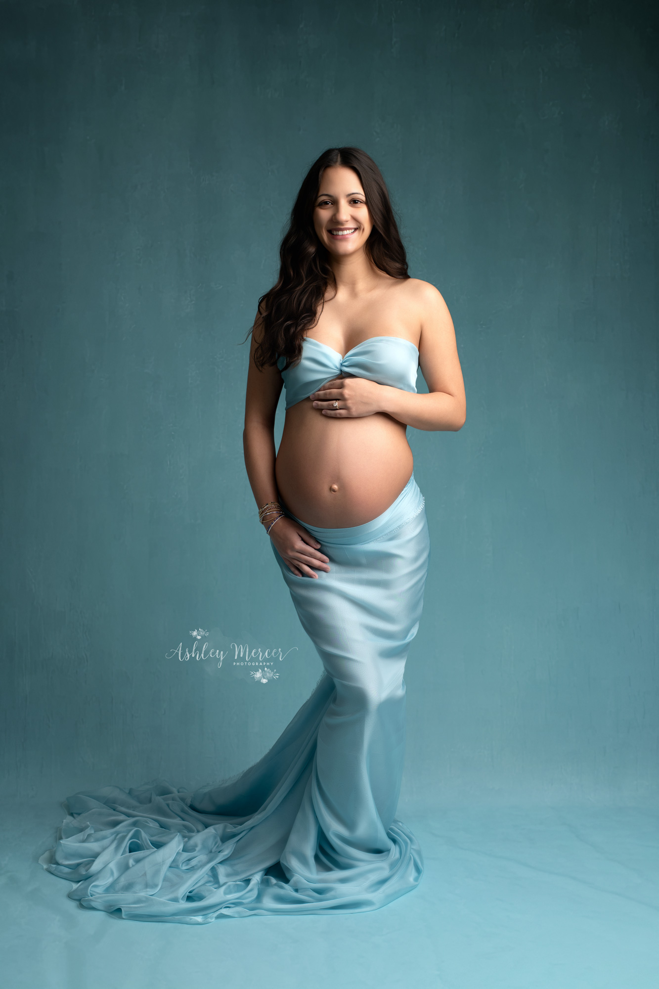 Smiling pregnant woman wearing a bandeau top and a matching mermaid style bottom is smiling while showing off her bare belly