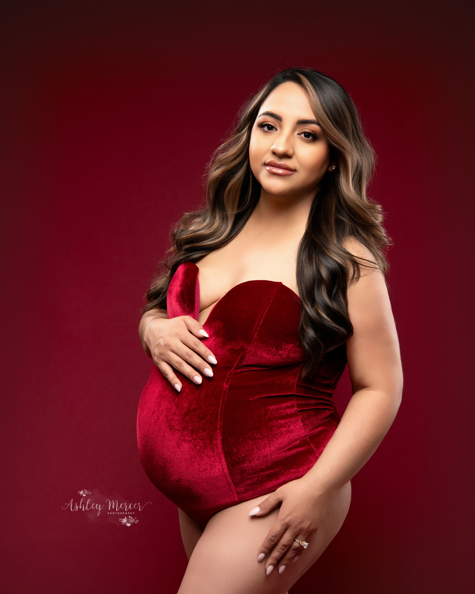 A three quarter cropped images of a pregnant woman holding her belly and looking directly into the camera. She is wearing a red velvet bodysuit with a dark red background giving a very fun and sexy feel.