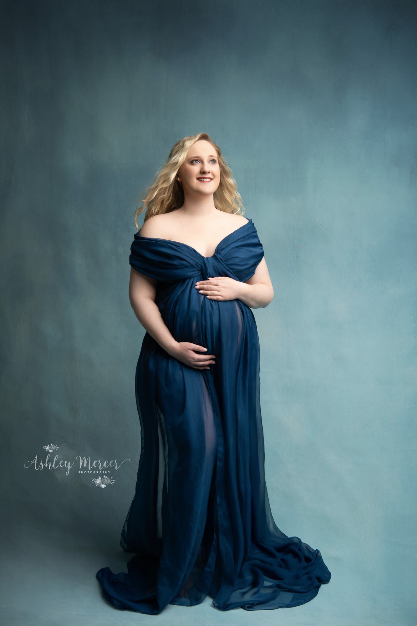 A studio maternity session with a light blue background and pregnant mom wearing a navy blue, off the shoulder, flowing gown. She is holding her belling and lovingly looking off into the distance.