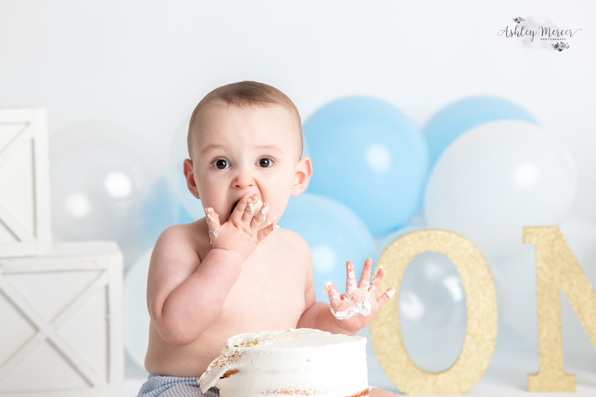 One year old birthday boy looking surprised as he puts a fist full of cake into his mouth