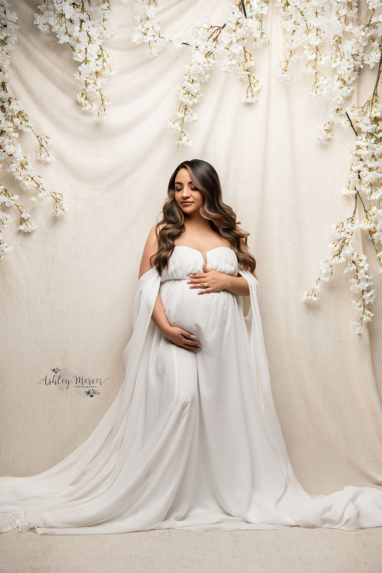 An expecting mother gently holding her belly with her eyes closed while against a wall of flowers.