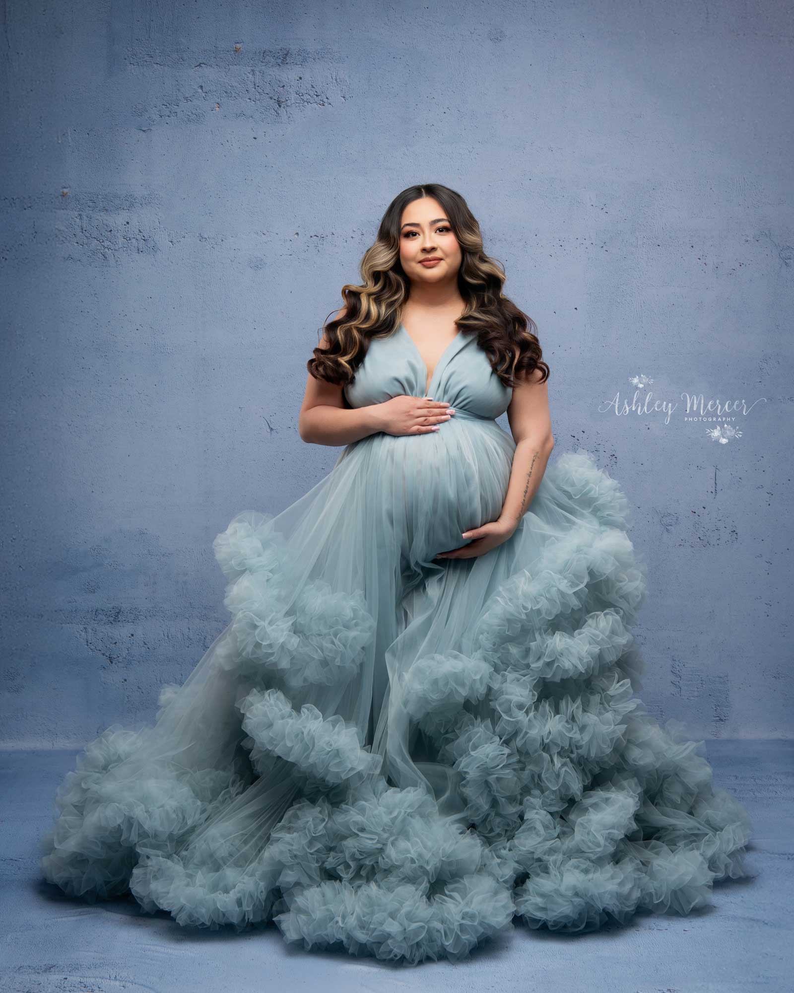 A monochromatic studio image with shades of blue. The pregnant woman is wearing a blue tulle gown, on a blue background, while looking at the camera