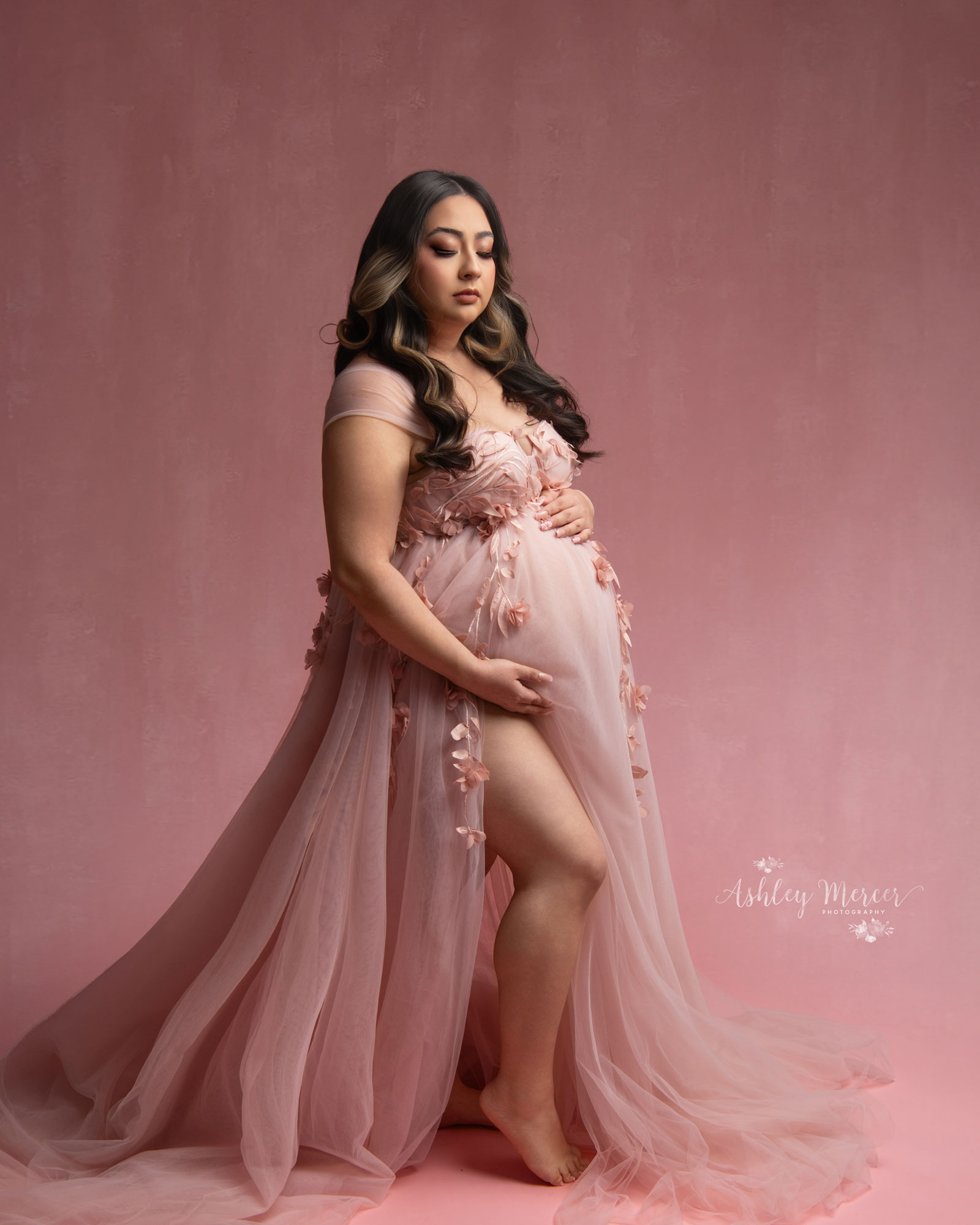 A studio maternity session with all pinks, pink background and a pink gown. The mom is posing with one hand under her stomach while looking down.
