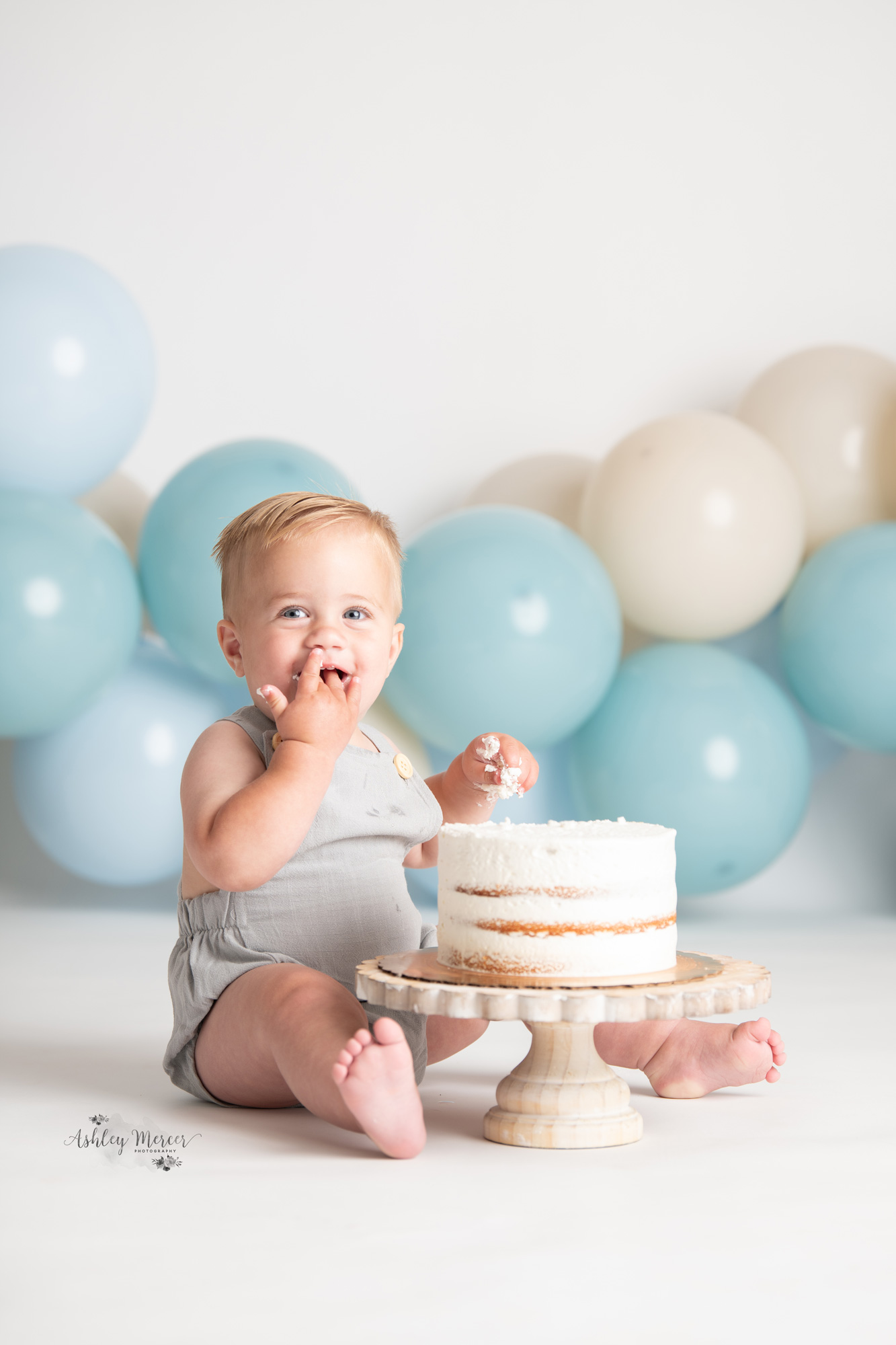 A cute one year old boy licking his fingers while eating his cake during a cakeshmash session.