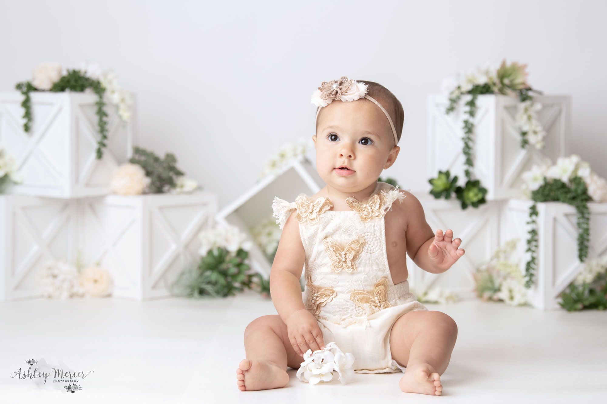 A one year old little girl wearing a white romper with butterflies is smiling at the camera.