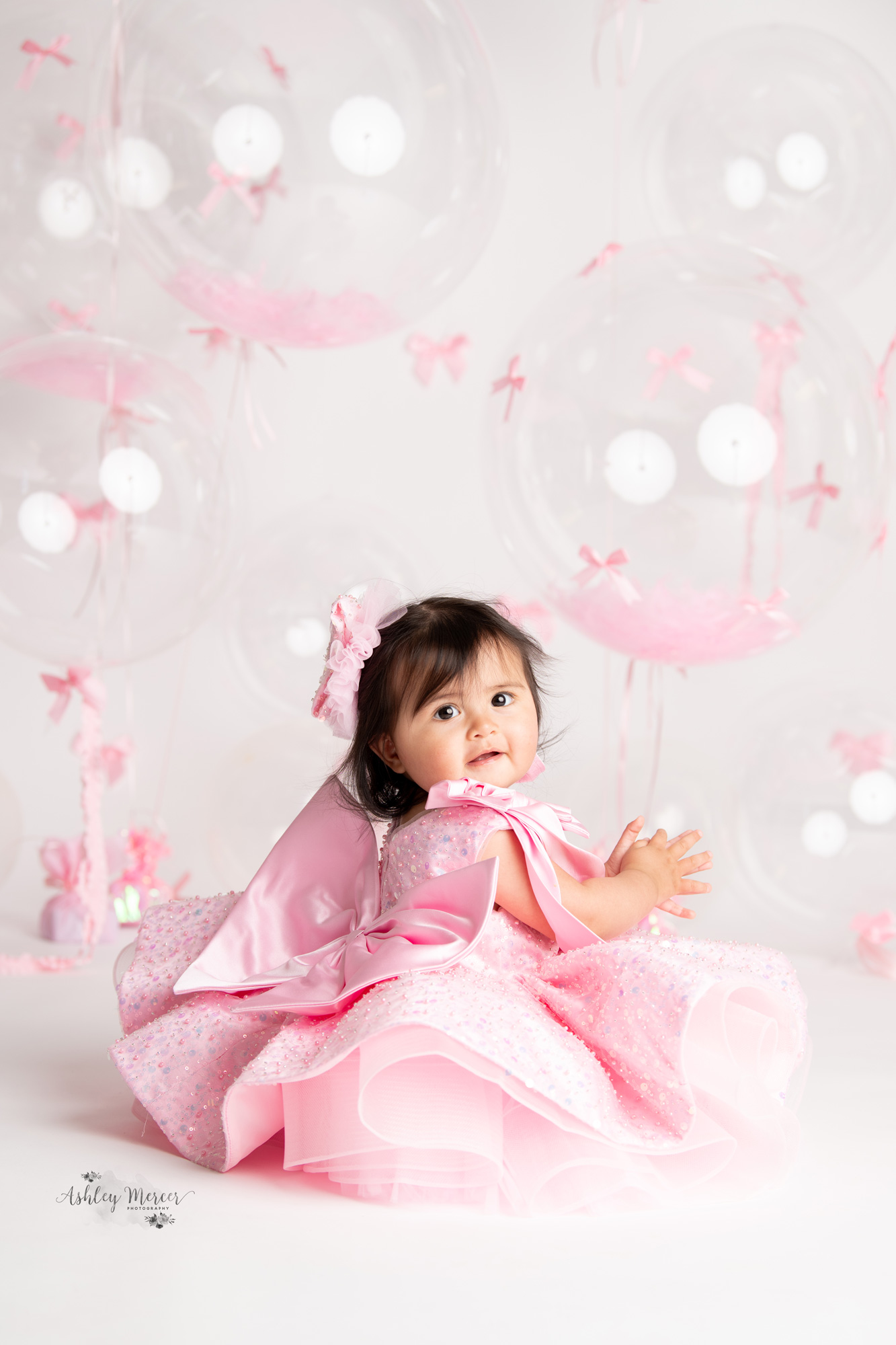 A one year old girl wearing a princess style dress looking over her shoulder at the camera. She is surrounded by by large balloons covered in small pink bows