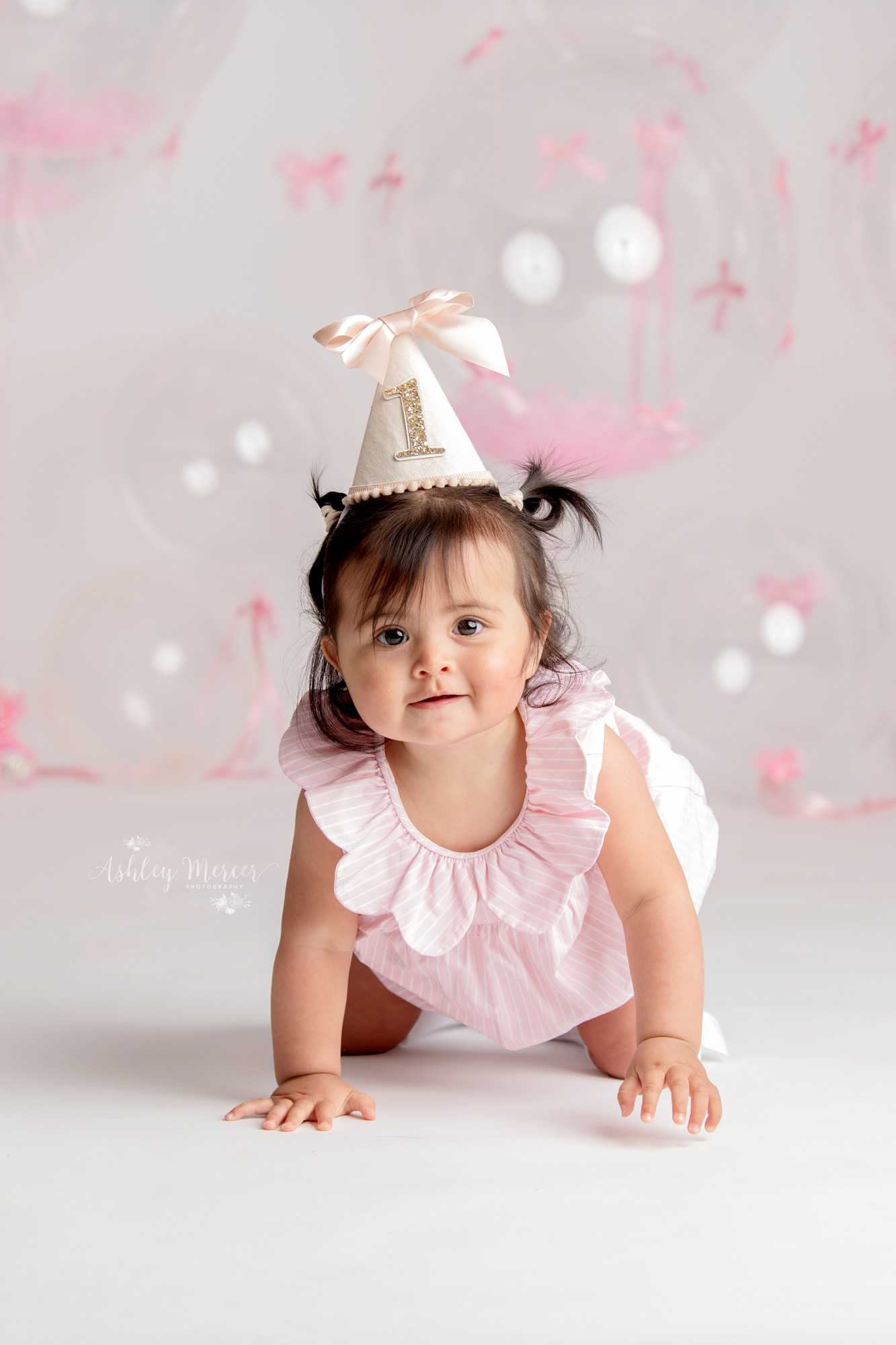 A happy one year old girl wear a 1st birthday party hat with lots of balloons and pink bows in the background