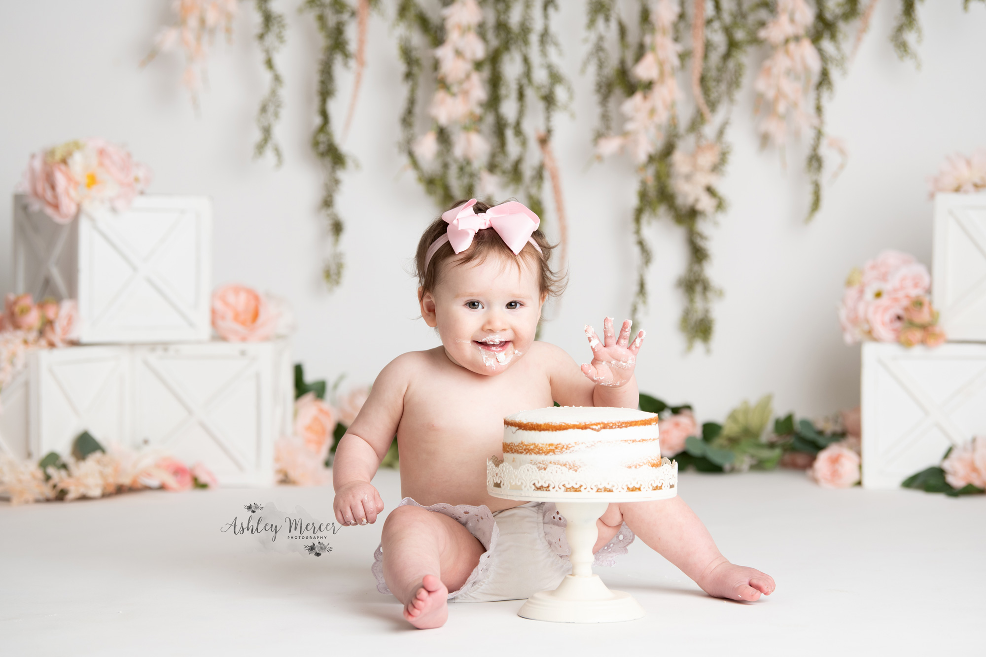 Little one year old girl wearing a pink bow is waving at the camera right before she digs into her first birthday cake. She is surrounded by light pink flowers and vines of greenery