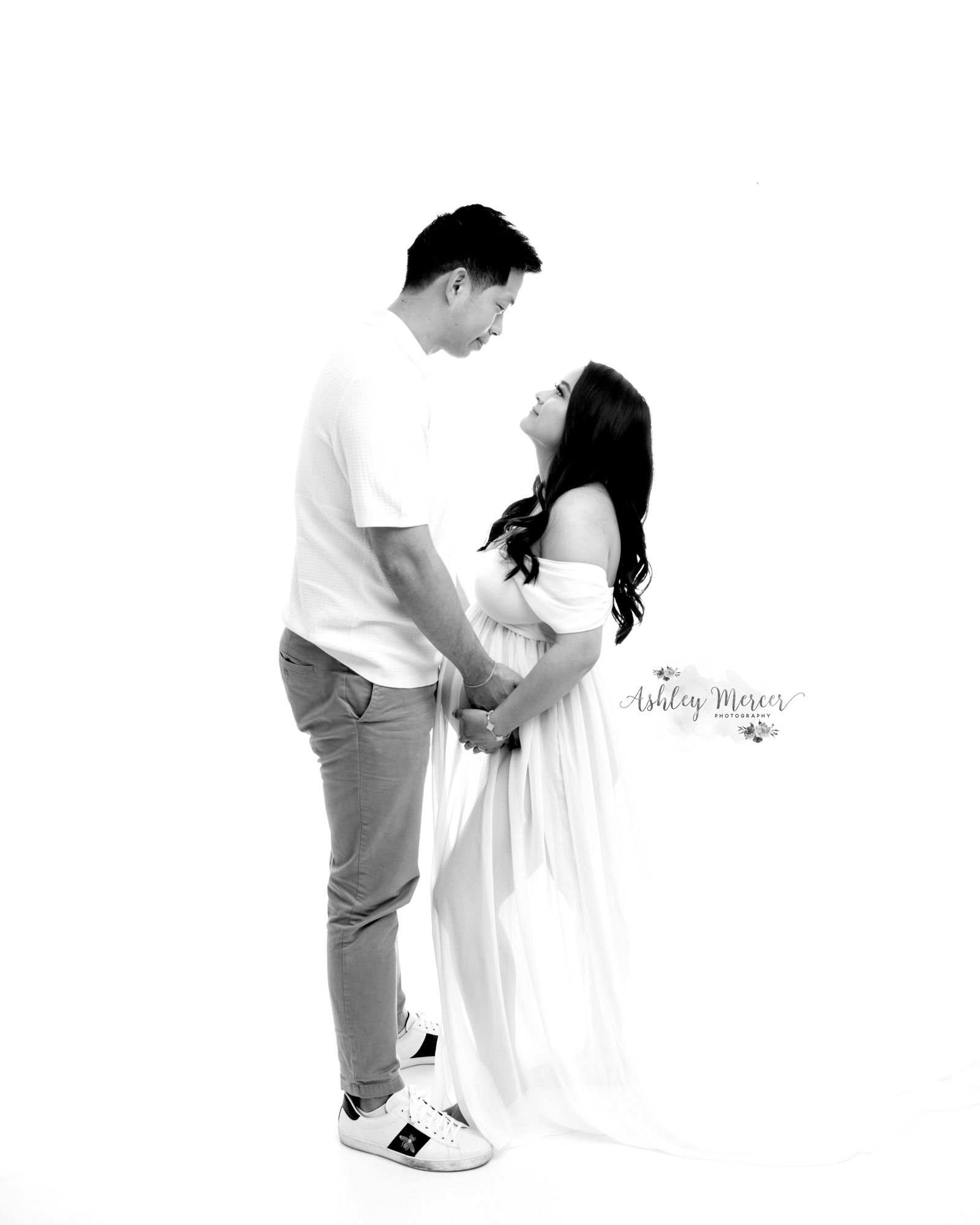 Timeless black and white studio image of a pregnant couple looking at each other. The dad and mom are both cradling her belly