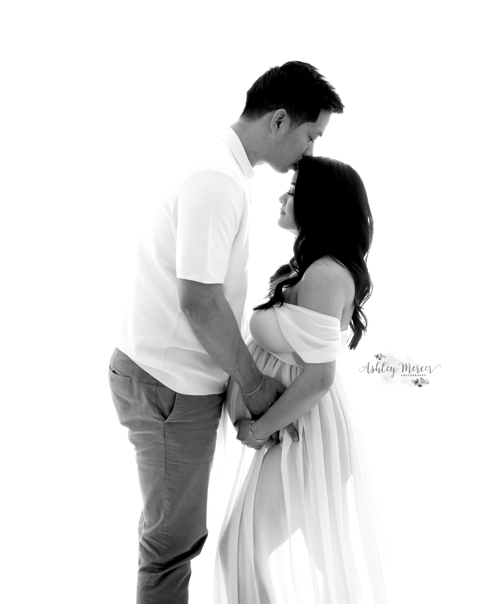 An elegant black and white studio image of a couple expecting their first child. The father is lovingly giving the mom a kiss on the head